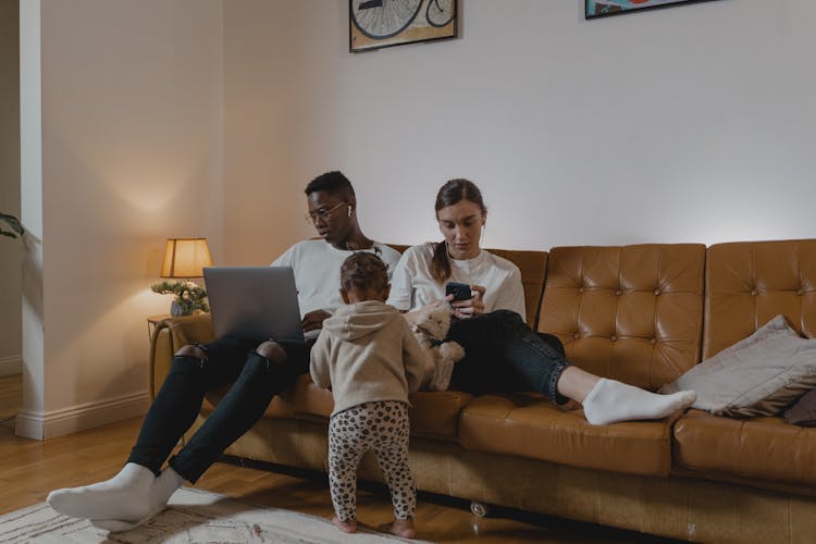 A Baby Standing Beside Couple Sitting On Couch Using Electronic Devices