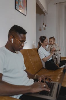 A man working on a laptop while a woman holds a baby on a couch, multitasking family life.