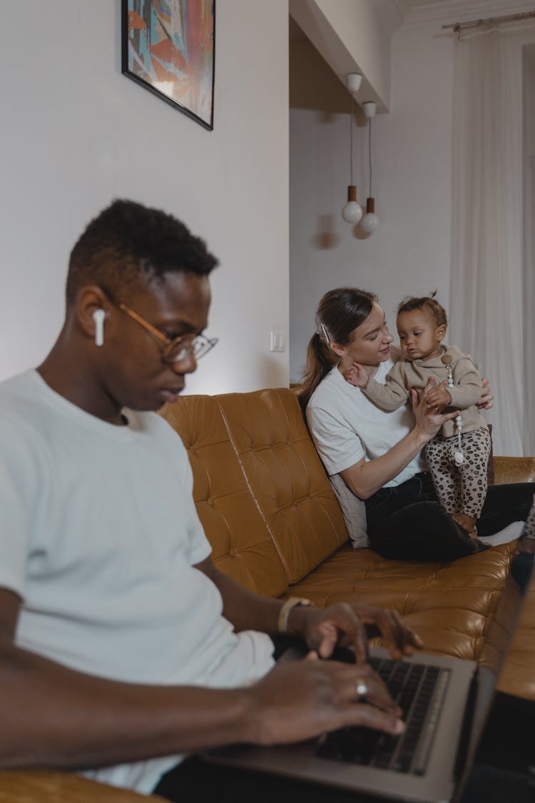 A Man In White Crew Neck Shirt Sitting On Couch Near A Lady Holding A Baby