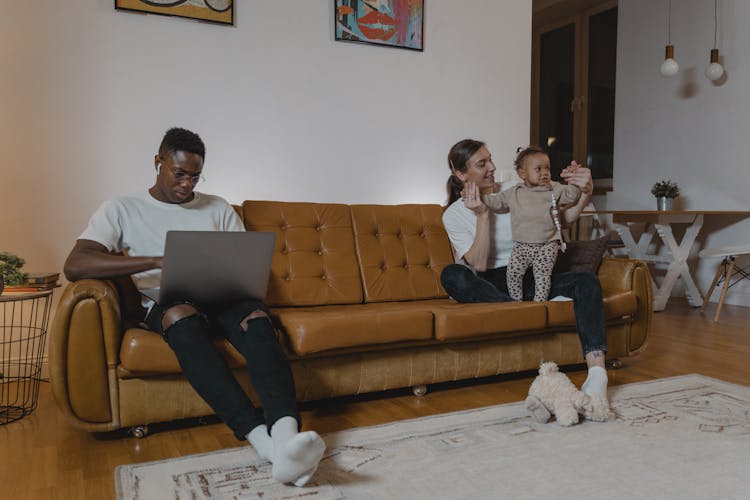 A Man In White Crew Neck Shirt Sitting On Couch Near A Lady Holding A Baby