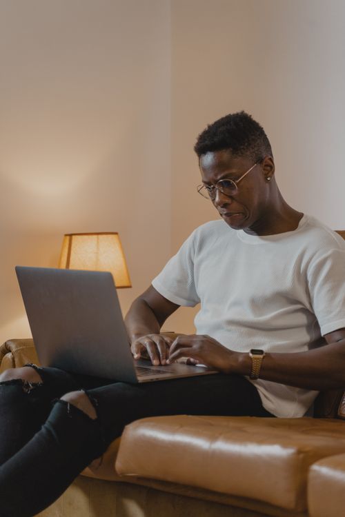 Free Young adult working on a laptop in a cozy living room setting with warm lighting. Stock Photo