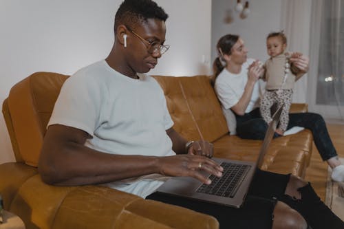 Man in White Crew Neck T-shirt Sitting on Brown Sofa while using Laptop