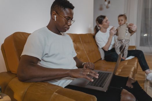 A man works on a laptop while a woman cares for a toddler on a sofa, highlighting multitasking at home.