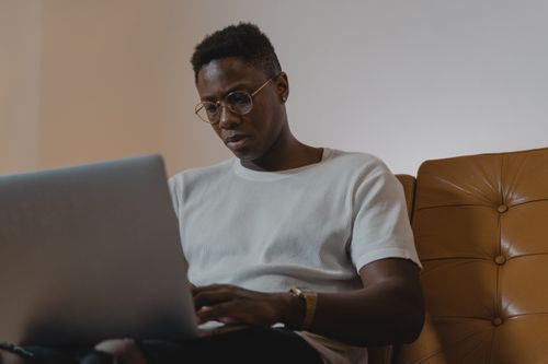 Free African American man focused on remote work from home using a laptop on a leather sofa. Stock Photo