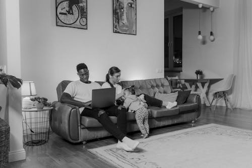 Grayscale Photo of a Man Using a Laptop next to his Wife and Daughter on a Couch