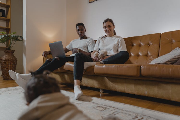 A Man And  A Woman Sitting On A Couch While Looking At The Baby Crawling On The Floor