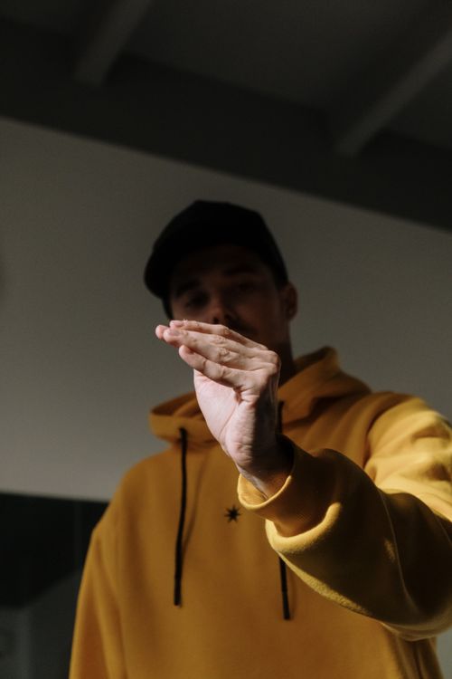Free A man in a yellow hoodie making a distinct hand gesture inside a dimly lit room. Stock Photo