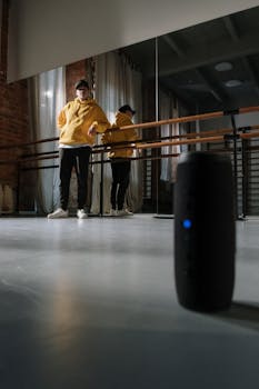 A man in a yellow hoodie stands in a dance studio next to a Bluetooth speaker.