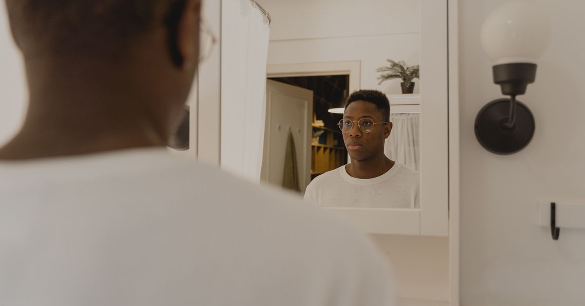 Um homem de óculos reflete seriamente em um espelho de banheiro, vestindo uma camisa branca.