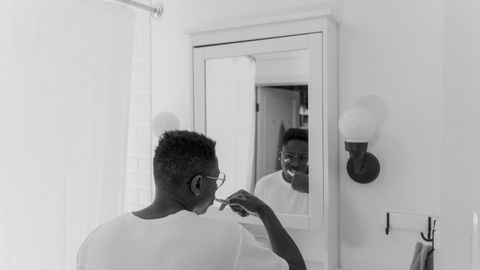 A black and white photo of a man brushing his teeth in front of a bathroom mirror.