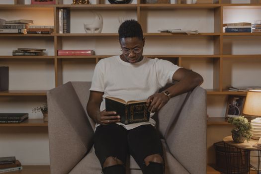 Young African American man with glasses reading a book in a cozy home setting.