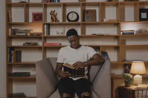 Free A man reading comfortably on a chair, surrounded by bookshelves in a cozy home library. Stock Photo