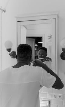 Black and white photo of man practicing oral hygiene, reflected in bathroom mirror.