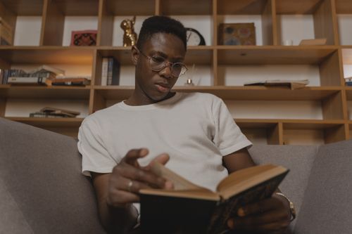 Free African American man reading a book in a comfortable modern indoor space with a stylish bookshelf. Stock Photo