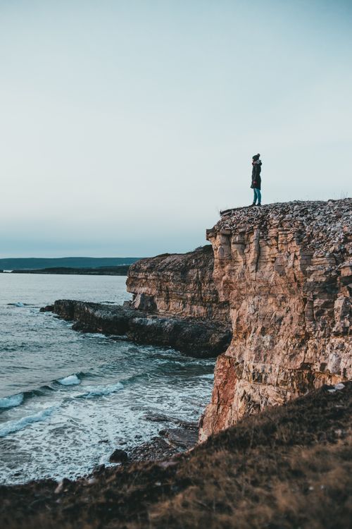 Free Side view of anonymous female tourist contemplating wavy ocean from rough cliff under light sky in cold weather Stock Photo