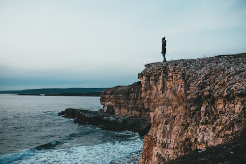 Unrecognizable woman standing on rocky cliff at seaside under cloudy sky