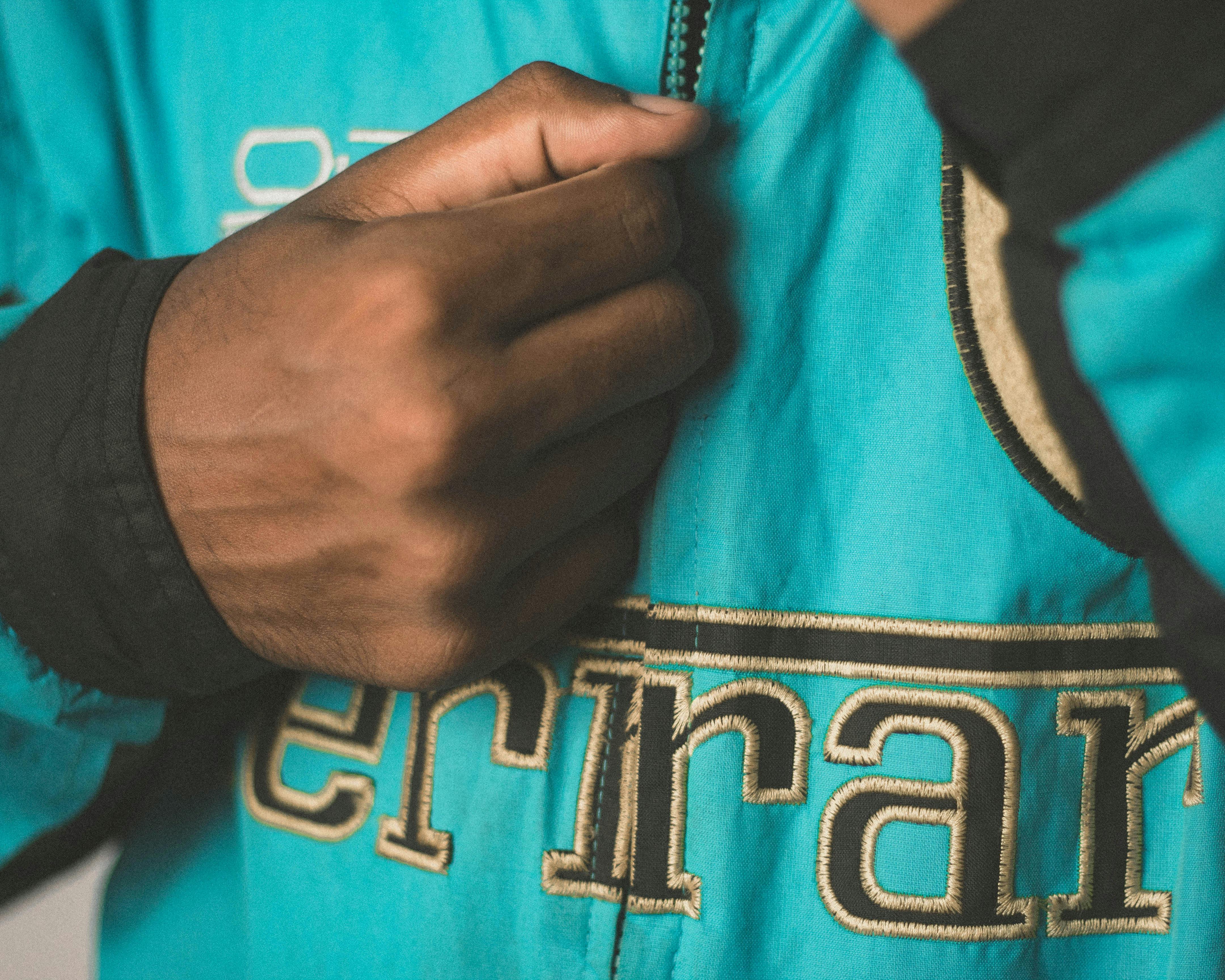 A close-up of a hand holding the zipper of a blue jacket with embroidered text.
