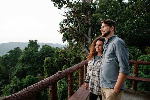 Free Sid view of cheerful young male and female travelers in casual clothes hugging and smiling while admiring picturesque nature from terrace during romantic trip in highland Stock Photo