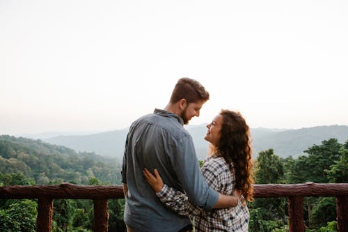 Back view of happy young romantic couple cuddling and looking at each other while standing on terrace against picturesque lush forest in mountains