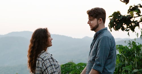 Side view of beloved young man and woman in casual clothes smiling and looking at each other during trip in green highlands
