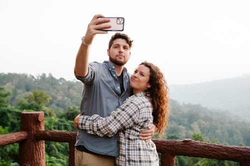 Joyful young romantic couple in casual shirts embracing while taking self portrait on smartphone standing on wooden viewpoint in mountains