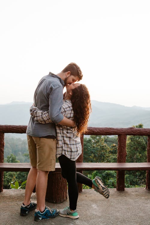 Free Side view of stylish young travelling couple in casual clothes hugging and kissing on observation deck in mountainous valley Stock Photo