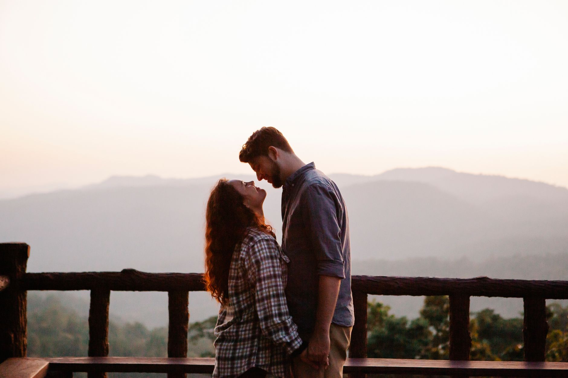 Side view of loving young casual couple holding hands and looking at each other while resting on terrace in mountainous valley at sunset