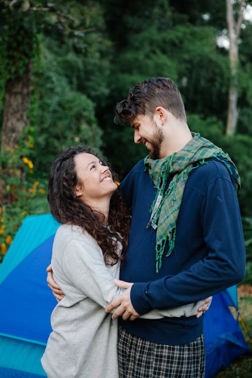 Happy couple smiling and embracing while looking at each other near blue tent among green trees in forest