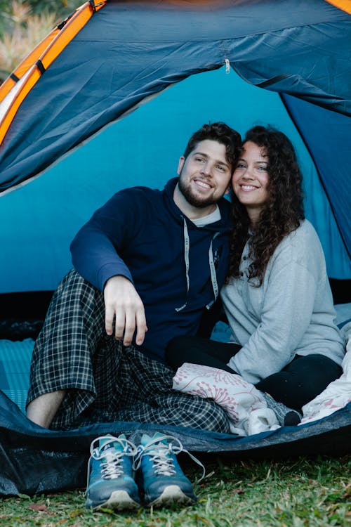 Positive young couple in casual outfit smiling and resting in tent on fresh green grass in nature