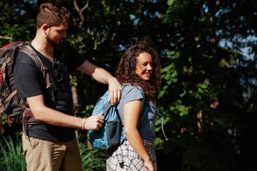 Side view of young bearded male hiker closing rucksack of cheerful female beloved against green trees