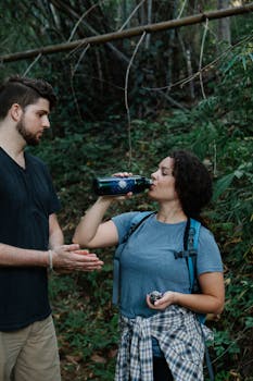Young couple of hikers in activewear with backpacks standing in forest and drinking water from bottle