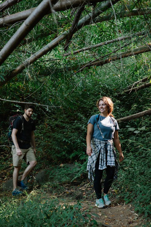 Full body of active young couple in casual clothes and backpacks smiling while walking on narrow pathway in lush green forest during trekking