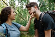 Side view of happy young woman smiling and touching face of tired boyfriend making grimace while standing together in green forest during hiking trip