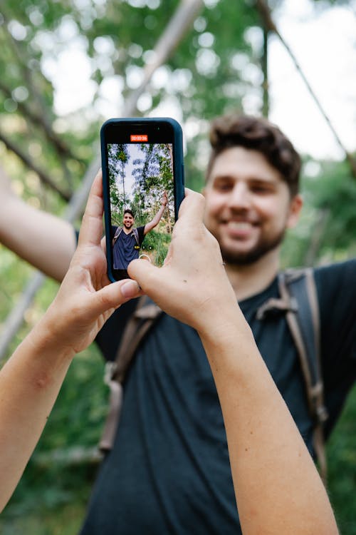 Free Crop unrecognizable woman taking photo of cheerful boyfriend on mobile phone while resting together in lush green jungle during summer vacation Stock Photo
