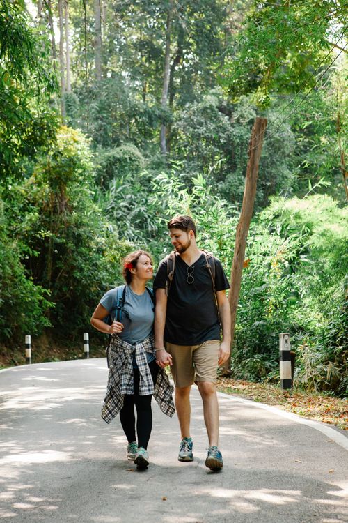Free Full body of positive young loving couple holding hands and looking at camera while walking along asphalt road near amidst lush green tropical trees during summer holidays Stock Photo