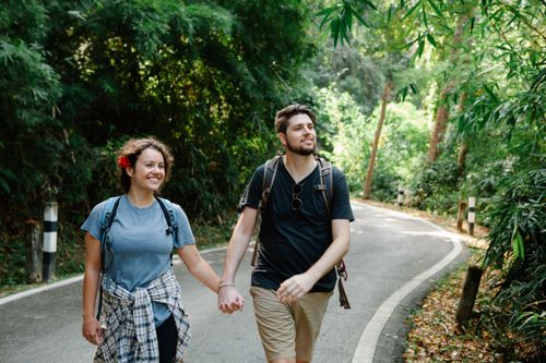 Free Positive loving young couple in casual outfits and backpacks holding hands while strolling on asphalt road between lush tropical trees during hiking in park Stock Photo