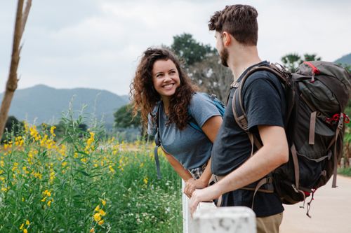 Free Happy young couple of tourists in casual clothes with backpacks looking at each other while leaning on wooden fence near grassy field with wildflowers in daytime near mountains and trees Stock Photo