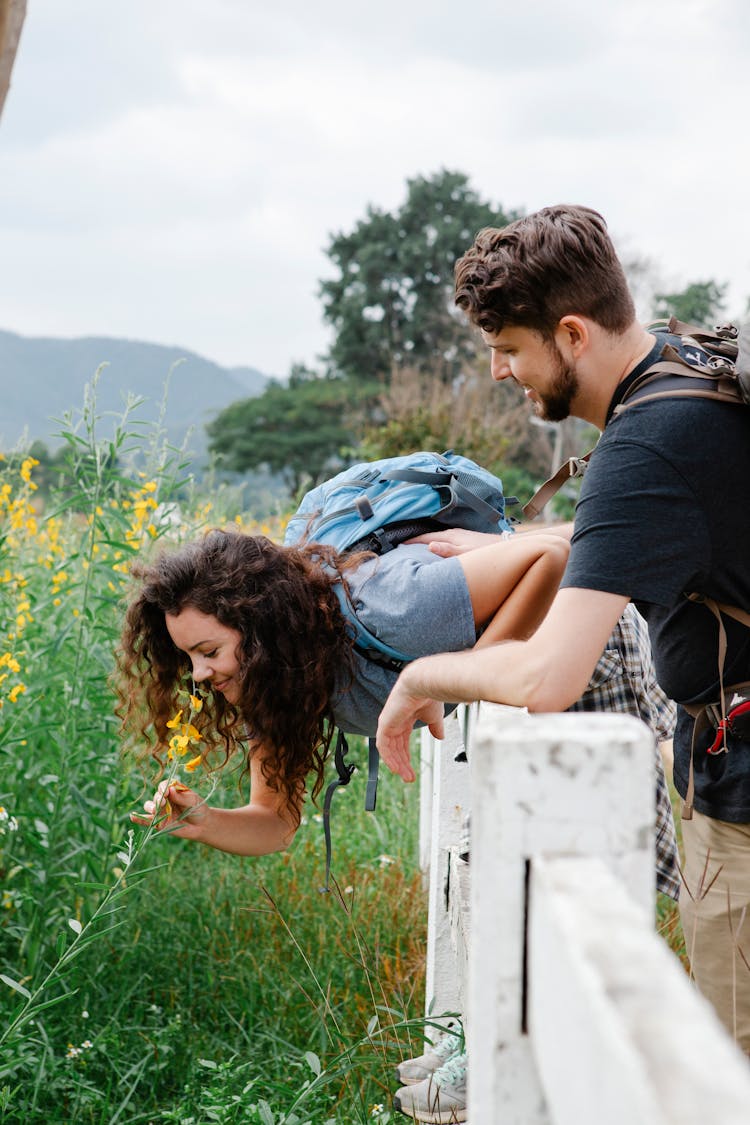 Couple Of Tourists On Fence Near Grassy Field With Wildflowers