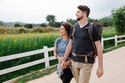 Free Young couple of travelers wearing casual clothes and backpacks while strolling on road near wooden fence and green grassy field with wildflowers near trees and looking away while holding hands Stock Photo