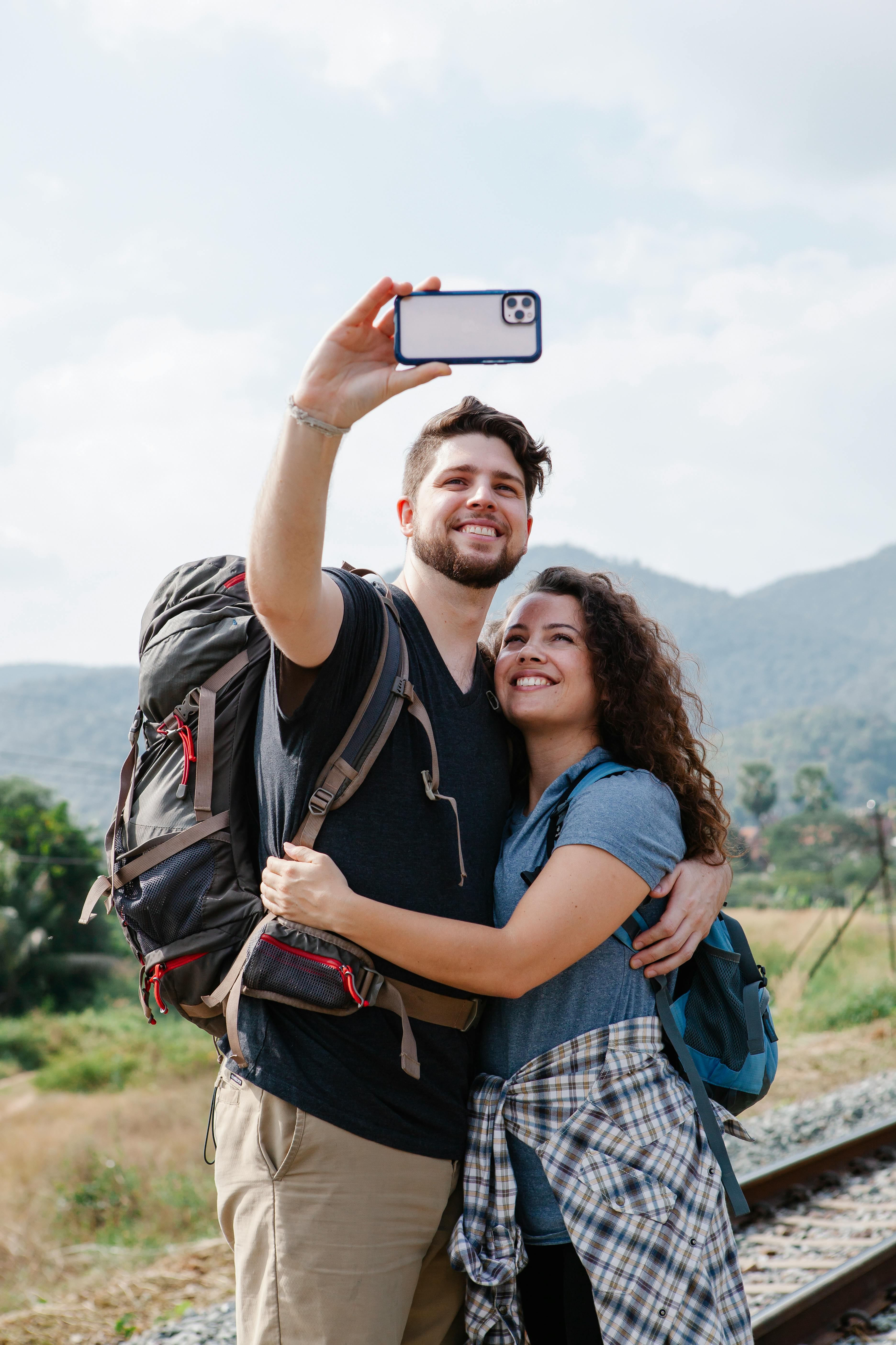 Smiling young couple of tourists in casual outfits with backpacks taking selfie on phone while standing and hugging near railway and grassy field with plants and hills in summer day