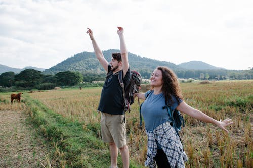 Young couple of travelers wearing backpacks and casual outfits with raised hands standing on field with dry and green grass with brown cow near trees and hills in summer day