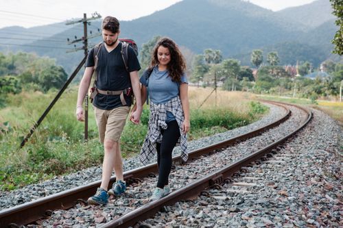 Free Cheerful couple of travelers in casual clothes with backpacks holding hands and  walking along railway placed in countryside against green mountains in daytime Stock Photo