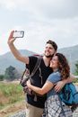 Happy couple of hikers in casual clothes with backpacks hugging and taking selfie on smartphone in nature against mountains in sunny day