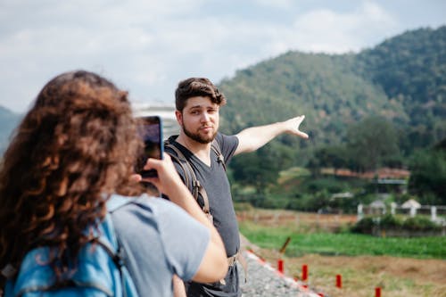 Free Back view of unrecognizable woman in casual clothes taking photo on smartphone of pensive boyfriend pointing at green hills during hike in countryside in sunny day Stock Photo
