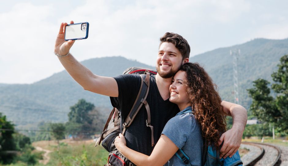 Cheerful couple of tourists in casual clothes with hiking backpacks hugging and taking selfie on smartphone in nature against green hills in daytime