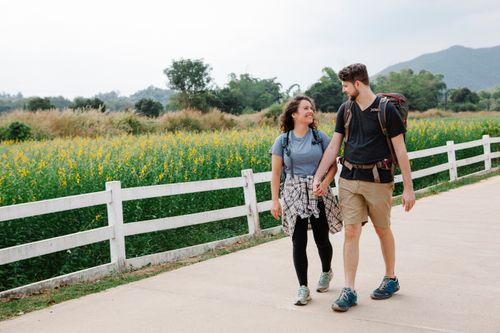 Free Cheerful couple in casual clothes holding hands and looking at each other while walking together on pathway near blooming field in countryside in daytime Stock Photo