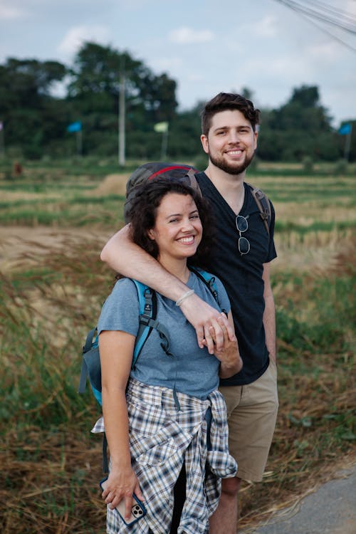 Positive couple of tourists in casual clothes with backpacks standing and embracing against green field in countryside in daytime