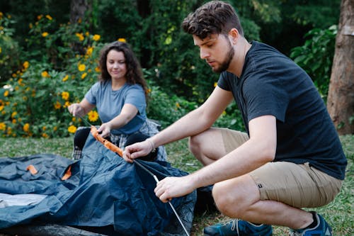 Concentrated couple of travelers in casual clothes squatting down and pitching tent in green forest during hike in daytime