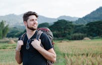 Positive man with trekking backpack standing in field against hills