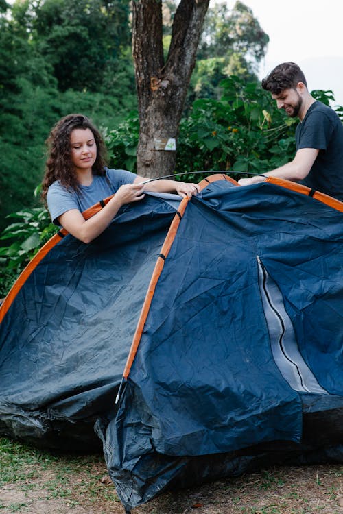Cheerful couple of travelers setting up tent during summer trip against trees in daytime
